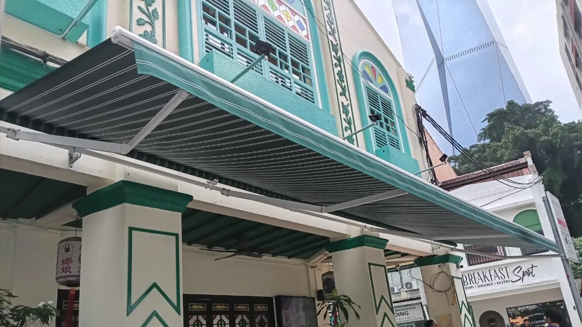 Looking up under a green striped awning at its white folding arms and the roller fixed to the wall